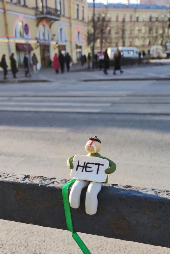 A figurine with a mustache sits on a fence holding a poster with the word NO, as people walk in the background.