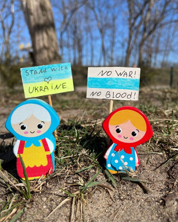 A doll set on grass holds an Ukrainian flag that reads, “Stand with Ukraine.” On its right, another doll holds a white and light blue flag that reads, “No war! No blood!”