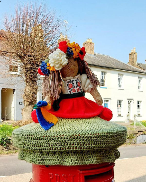 A multicolored woven doll holds a flag while sitting on a mailbox.