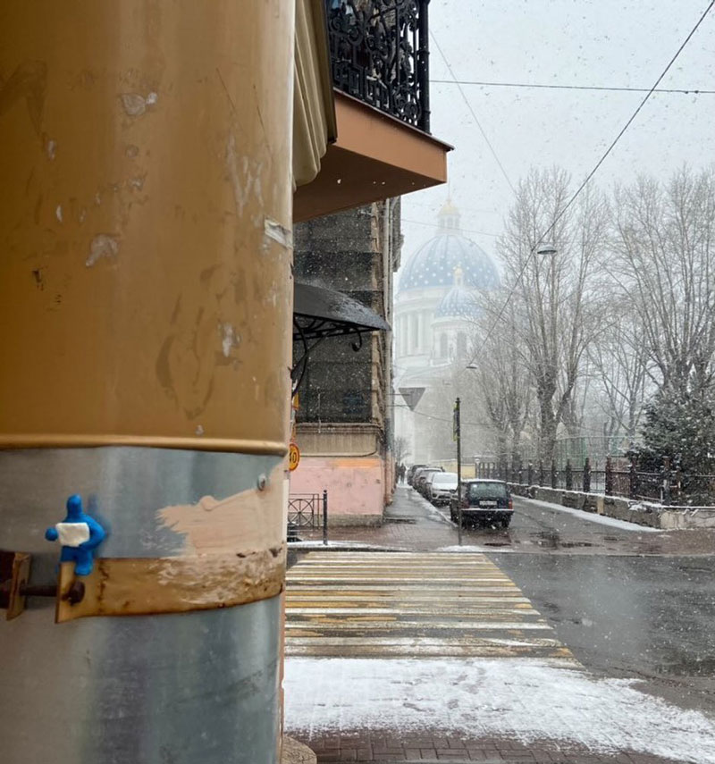 A blue figurine, seated on a post at the side of the street, holds up a blank poster. In the background is a snowy street.