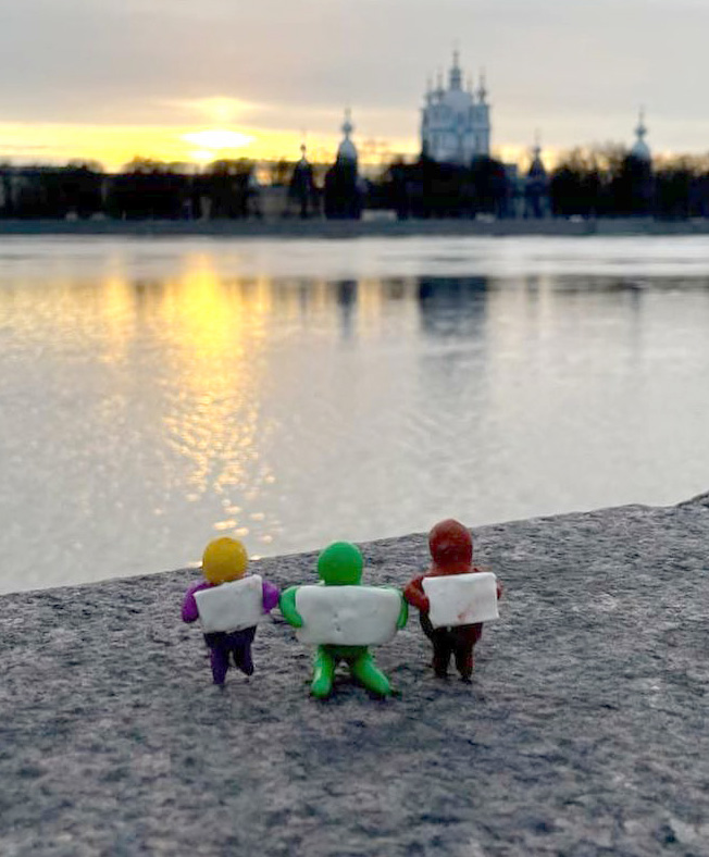 Three figurines hold blank posters by a river, a large church in the background.