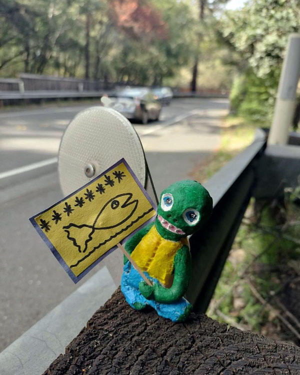 A green figurine holding a yellow poster with asterisks and a drawing of a fish.